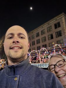 A parade float draped in Christmas lights rolls by as Sam and I flip the camera for a selfie. Behind the float is one of Modesto's historic buildings, a four-story brick building with a grid of windows. The night sky is lit by the moon, blurred by light clouds. 