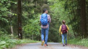 A father and daughter, wearing backpacks, head down a wide path into the woods.